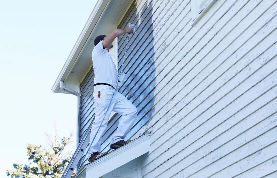 Painter on a ledge using a roller to apply fresh paint to the upper exterior of a weatherboard house.
