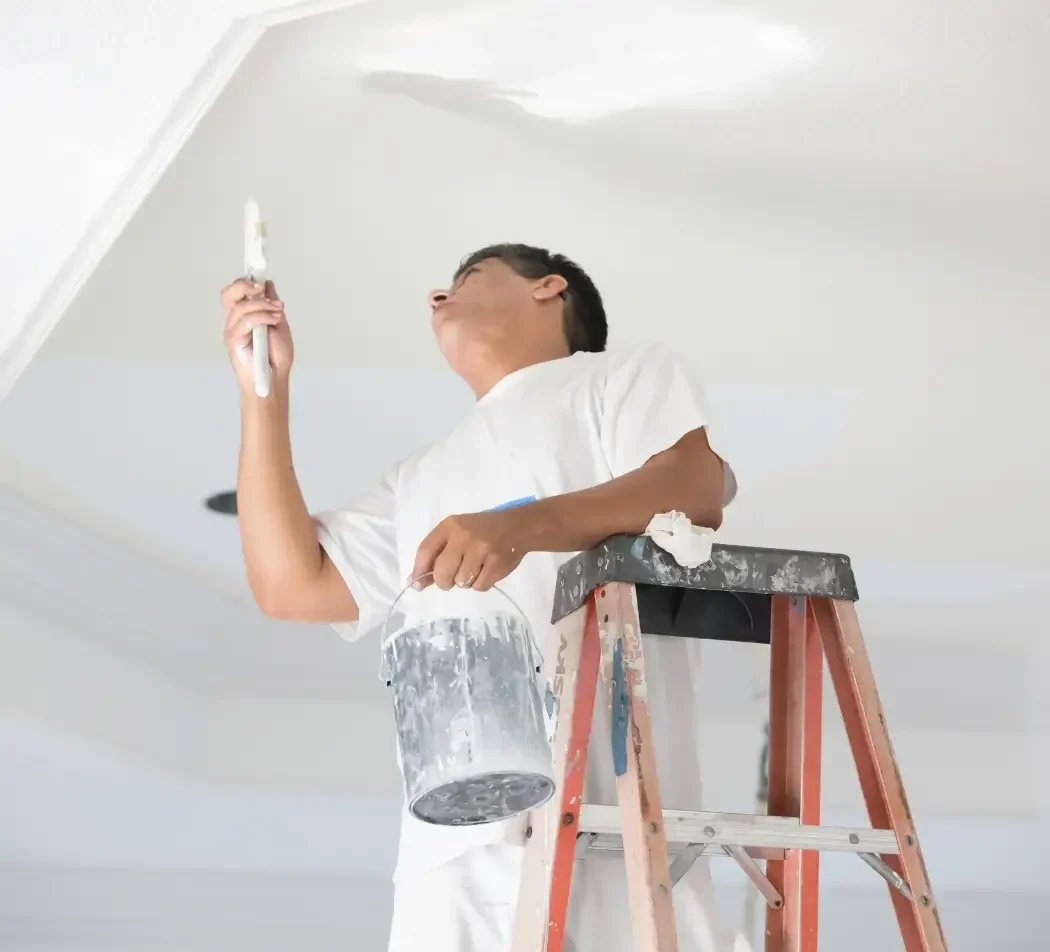 Painter on ladder touch-up painting the ceiling of a residential home with a brush and paint bucket